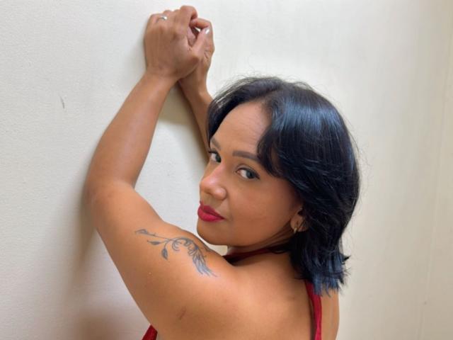 Loraine Clof, avec une peau brun moyen, des cheveux noirs mi-longs, du rouge à lèvres et un tatouage floral sur le haut de son bras, accoudée contre un mur blanc, les bras levés, regardant par-dessus son épaule, sexy..