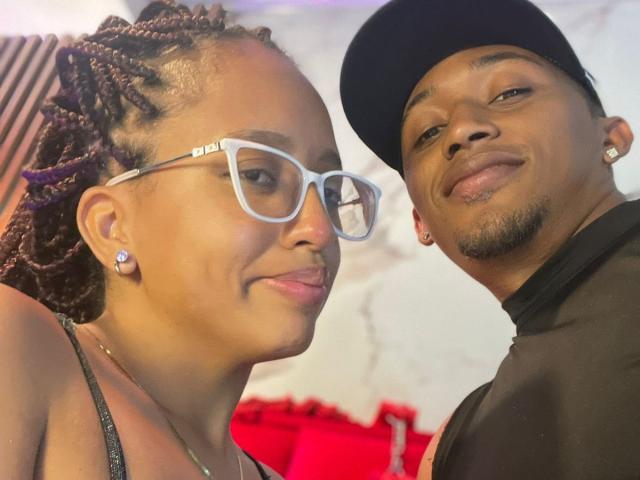 Close-up photo of LilithAndAsmodeo and another guy. LilithAndAsmodeo has braided hair, white glasses, earrings, sticking tongue out, looking sexy. Guy has black cap, goatee, black shirt, looking smug. Red background.