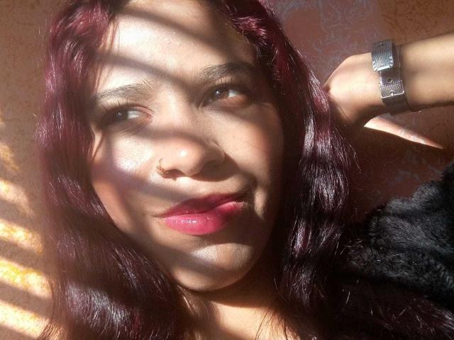 Close-up of ValerieWoods with dark red hair, sunlight casting shadows on her face, wearing a black top, red lipstick, silver ring on her right hand, slightly smiling, looking seductive.