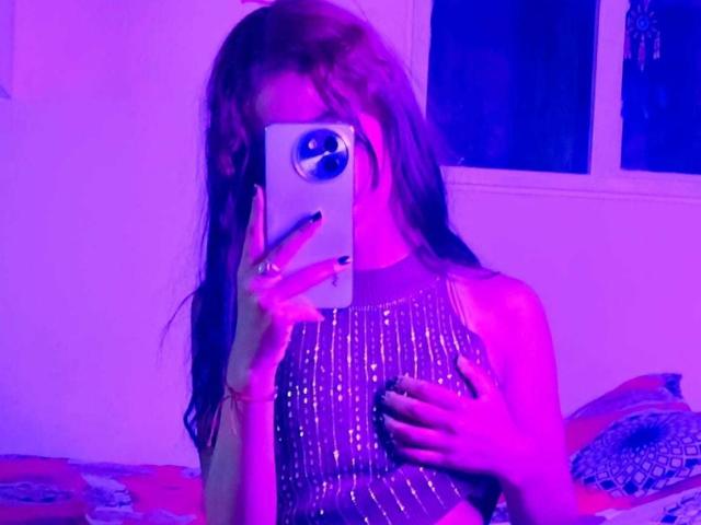 Une photo de ValerieWoods, prise par elle-même, où elle a de longs cheveux bruns, tient un téléphone devant son visage et porte une tenue sans manches scintillante. le torse, éclairé par des lumières violettes et bleues, paraissant séduisant.