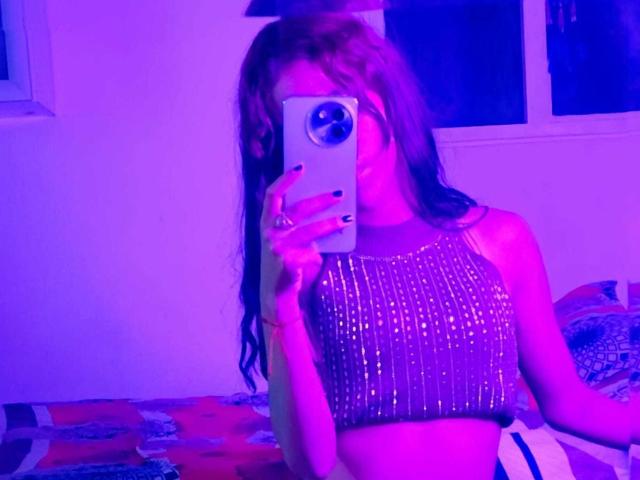 ValerieWoods se prend un selfie devant un miroir, éclairée par une lumière néon violette-bleue. Elle porte un haut scintillant sans manches, tient son téléphone d'une main et on peut voir ses longs cheveux noirs. Le décor se situe dans une pièce faiblement éclairée..