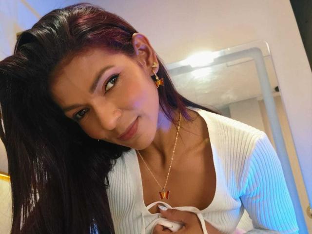 Close-up of HannaPretty with dark, straight hair leaning forward, smiling seductively. She's wearing a white, ribbed, low-cut top, gold necklace with a small pendant, and gold earrings. Bright lighting.