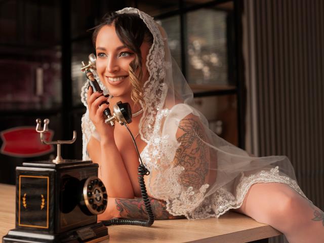 AnastasiaVolkov, com tatuagens no braço, está deitada em uma mesa, vestindo um véu de noiva branco rendado e lingerie, segurando um telefone antigo, com uma expressão sedutora e sorridente, criando tensão sexual. Telefone antigo em primeiro plano.