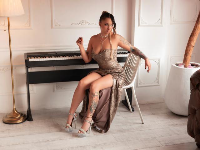 Anastasia Volkov, in einem glitzernden, einarmigen Kleid, sitzt auf einem Stuhl, die Beine gekreuzt, und zeigt ihren tätowierten Oberschenkel. Sie trägt High Heels, die Hände sind erhoben, eine sinnliche Spannung liegt in der Luft. Im Hintergrund stehen ein Klavier und eine Lampe..