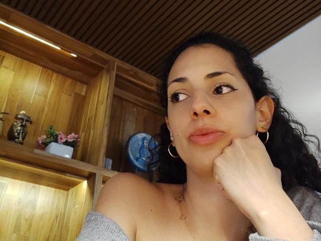 Close-up photo of CurlyBlossom, a woman with curly black hair, light brown skin, and hoop earrings, resting her cheek on her hand, looking to the side. Wooden shelves in background.