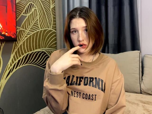 VeolaVanhoecke, une femme blanche aux cheveux bruns mi-longs, mordille son doigt tout en portant un t-shirt beige avec l'inscription "California West". Sweat-shirt "Coast", avec un aspect séduisant, avec une lumière rouge néon en arrière-plan..