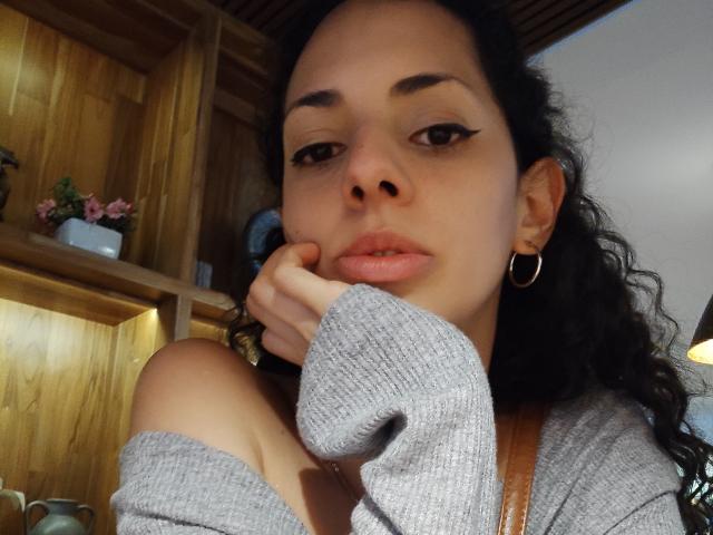 Fotografía de primer plano de CurlyBlossom, una mujer curvilínea con cabello oscuro y rizado, que lleva una blusa gris con un hombro descubierto, con una mano apoyada en mejilla, mirando a la cámara con los ojos entrecerrados, pequeños aros..