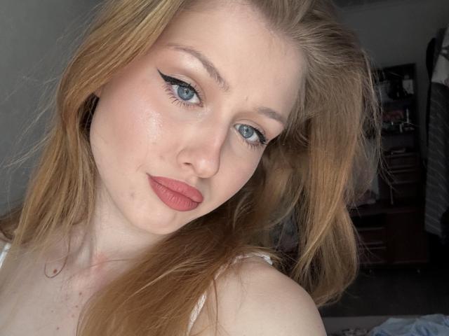 Fotografía de primer plano de Nikollia, una mujer de piel clara con cabello rojo, ojos azules y lápiz labial rojo, mirando directamente a la cámara con una ligera inclinación. La luz suave resalta su rostro. Increíblemente sexy..