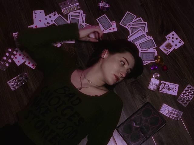 VeolaVanhoecke lies on a dark wooden floor surrounded by scattered playing cards and dice. She wears a black top, her expression is sultry, with intense eye contact and red lipstick.