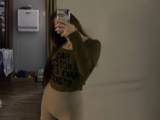 Fotografia di VeolaVanhoecke, selfie allo specchio, che tiene in mano il telefono, coprendosi il viso, indossa un maglione verde a coste con la scritta "TASH IS AN testo "INCREDIBILE", pantaloni beige a vita alta, in piedi di lato, stanza poco illuminata, porta e parete sullo sfondo..