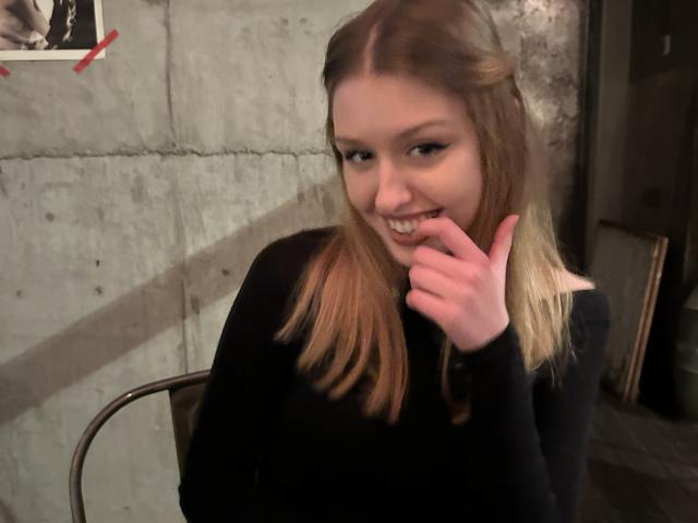 Nikollia, a young woman with fair skin and blonde hair, bites her finger while smiling seductively. She wears a black top, with a concrete wall and shadows in the background.