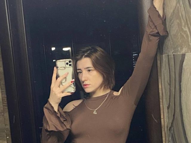 VeolaVanhoecke, a slim woman with short brown hair, takes a mirror selfie. She wears a brown long-sleeve top with cutouts, lifting her right arm. She has red nails and a necklace. Her expression is neutral.