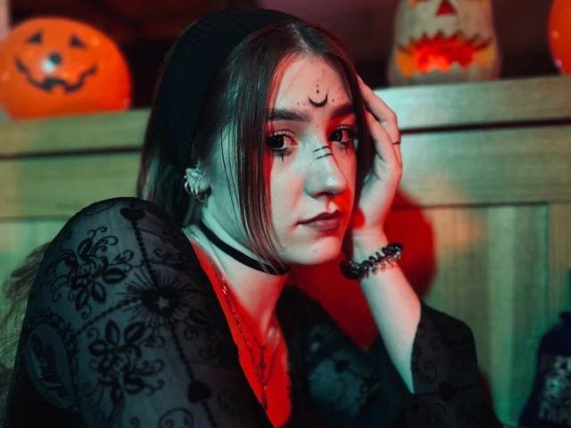 VeolaVanhoecke, donna asiatica con lunghi capelli neri, indossa un kimono nero con fiori, una camicia rossa, una collana nera e un braccialetto. Guardando seducentemente verso la fotocamera, con una mano sul viso, zucche di Halloween sullo sfondo..