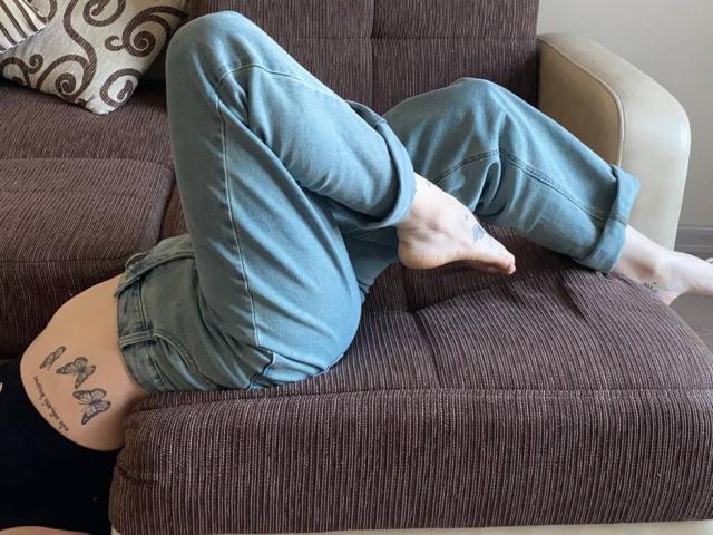 VeolaVanhoecke è sdraiata a faccia in giù su un divano marrone, con le gambe piegate sulle ginocchia, indossando dei jeans blu. Tatuaggio visibile sulla coscia destra, raffigurante tre farfalle. Piedi in vista, posizione rilassata e informale..