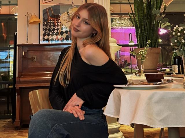 Nikollia with long, straight brown hair, wearing a black off-shoulder top and blue jeans, sitting at a table with a white cloth, looking back with a slight smirk. Fucking sexy vibe.