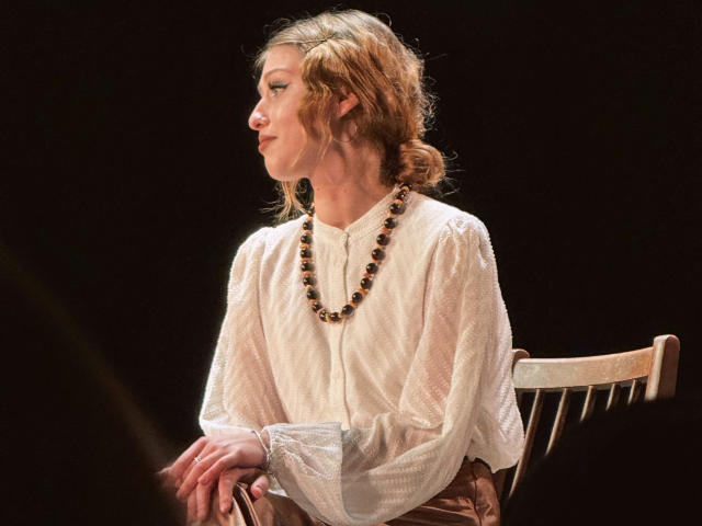 Nikollia sits sideways on a chair, wearing a white, textured blouse and a black bead necklace. Her brown hair is tied back. She has a focused expression, black background.