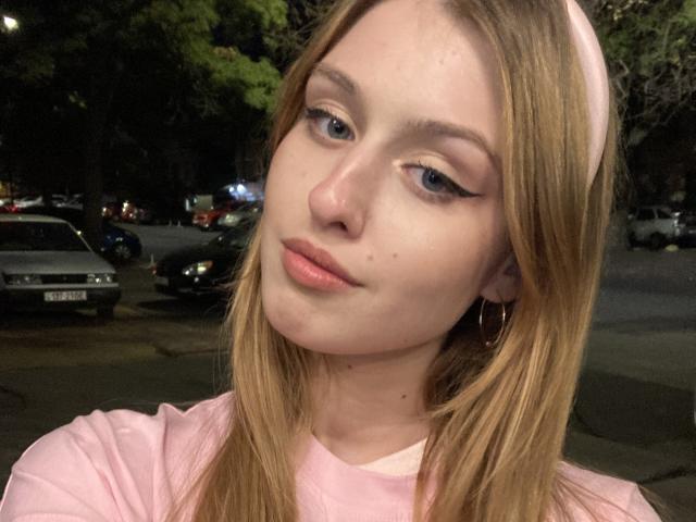 Close-up de Nikollia, uma jovem com pele clara e cabelo castanho claro, vestindo uma camisa rosa. Ela olha ligeiramente para o lado, com um sorriso discreto, e os olhos semicerrados. Fundo de rua urbana.