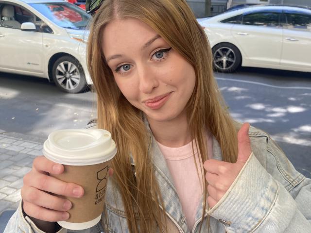 Nikollia, with long brown hair and blue eyes, holds a coffee cup in her right hand, giving a thumbs-up with her left. Wearing a denim jacket over a pink top, cars in background.