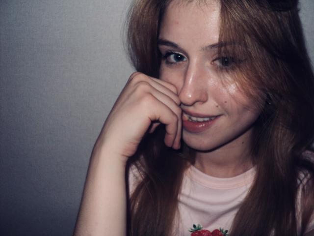 Close-up of Nikollia, a young woman with fair skin and long brown hair, resting her chin on her hand, gazing at the camera with a slight smile. She's wearing a white tank top.