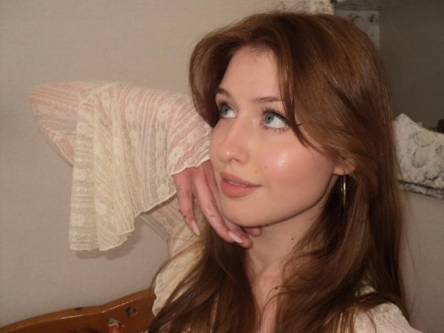 Nikollia, a brunette woman with long hair, gazes sideways with a slight smile. She rests her cheek on her hand, wearing a white, textured blouse. Soft lighting highlights her blue eyes.
