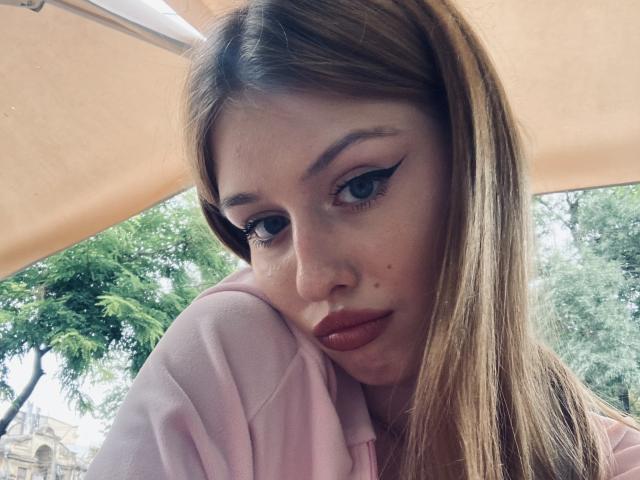 Close-up of Nikollia, a brunette woman with long hair, heavy eyeliner, and red lipstick, resting her head on her hand. She's wearing a pink shirt, with trees in the background. Fucking sexy.