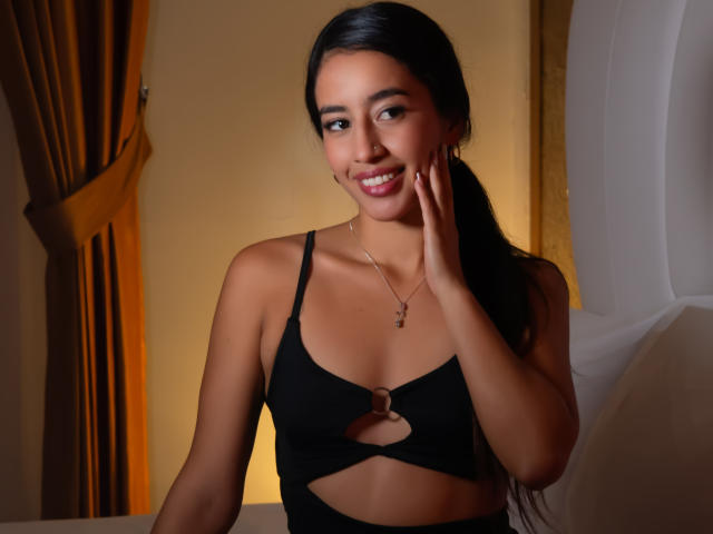 Fotografia di Alyssa Torres con lunghi capelli neri, che indossa un reggiseno nero con inserti aperti, sorridente e che si tocca il viso. Luce calda, tende marroni sullo sfondo. Tensione sessuale, aspetto attraente, espressione sicura di sé..
