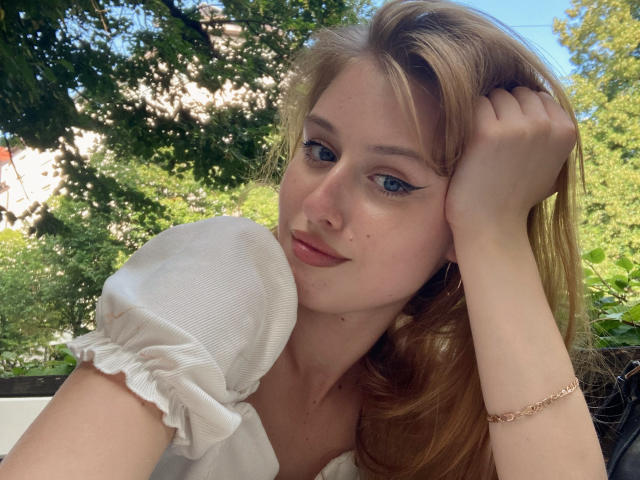 Foto van Nikollia met lang blond haar, blauwe ogen en lichte make-up, die een witte top met pofmouwen draagt, met haar hand in haar, subtiele glimlach, buiten, met groene bomen en zonlicht. Absoluut prachtig.