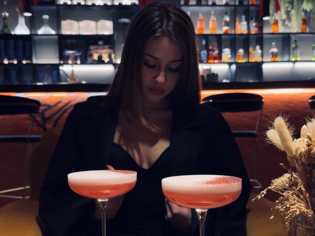 Milagros Edmisten, uma mulher de cabelos castanhos longos, está sentada em um bar, olhando para dois coquetéis rosa com espuma por cima. A iluminação suave e com tons escuros realça as bebidas e as prateleiras do bar ao fundo..
