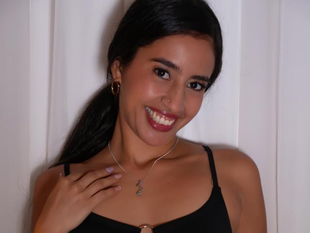 Fotografie von Alyssa Torres mit dunklem Haar im Pferdeschwanz, die lächelt und roten Lippenstift trägt, und die ein schwarzes Tanktop und eine goldene Halskette trägt. Kette, Hand in der Nähe der Brust, weißer Hintergrund, sexy und selbstbewusst..