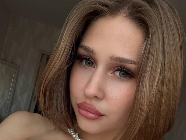 Close-up van Milagros Edmisten met glad bruin haar, volle lippen en donkere eyeliner. Ze kijkt recht in de camera met een lichtvoortreffende uitdrukking, en draagt een parelketting..