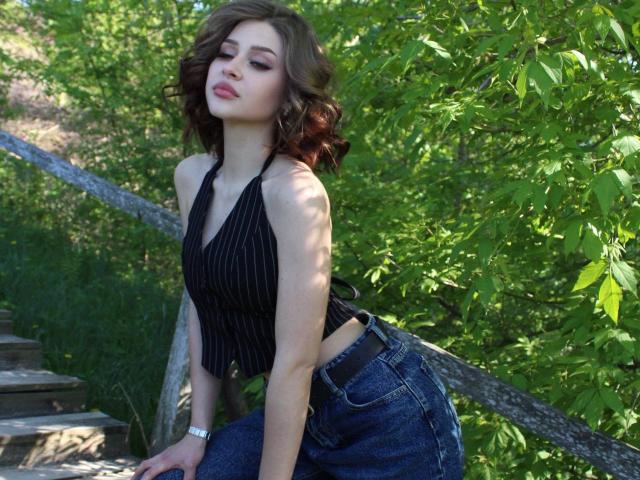 Milagros Edmisten, com cabelo castanho na altura dos ombros, vestindo um top preto com listras finas e calças jeans de cintura alta, está inclinada. apoiada em um corrimão de madeira, cercada por folhagem verde vibrante, olhos semicerrados, lábios levemente entreabertos.