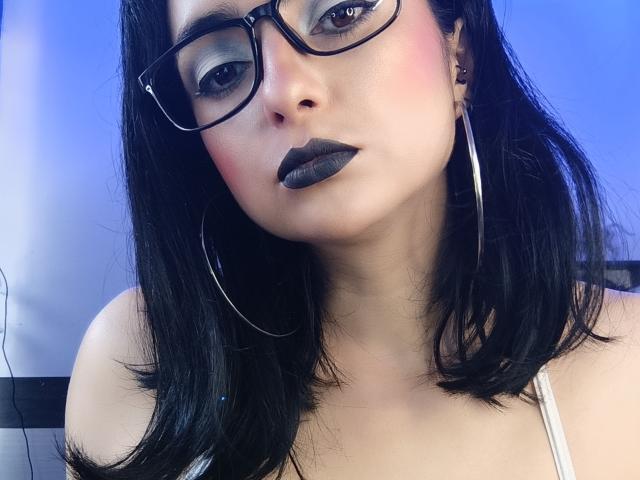 Close-up foto van CandyParis, een vrouw met zwart haar, zwarte lipstick en een bril, die recht in de camera kijkt. Ze draagt een witte top, grote oorringen, met een blauwe achtergrond. Extreem intense blik.