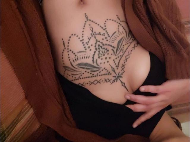 Spiceupyourlife-hot arbore un tatouage encre noire, très détaillé, représentant un motif floral sur le bas de son ventre. Portant un cardigan marron et des sous-vêtements noirs, avec une main posée sur leur ventre. Tension sexuelle.