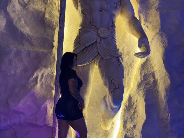 Silhouette de LindaAbler, aux formes généreuses, portant un short moulant, debout devant une statue musclée et angélique, fissurée et lumineuse. figure avec un laurier.
une lumière jaune vif contraste avec des ombres violettes. Tension sexuelle
.