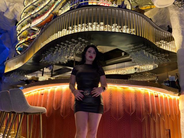 LindaAbler stands in a modern bar, wearing a sheer black dress that reveals her nipples and a belt with a gold buckle. Her hands rest on her hips, exuding confidence. Bright neon lights and a curved bar with hanging glasses form the background.