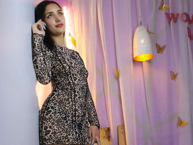 ArisSuarez staat leunend tegen een witte muur, ze draagt een strakke jurk met luipaardprint en houdt een telefoon op haar oor. Het warme licht van een hanglamp verlicht haar, omringd door vlinderdecoraties..