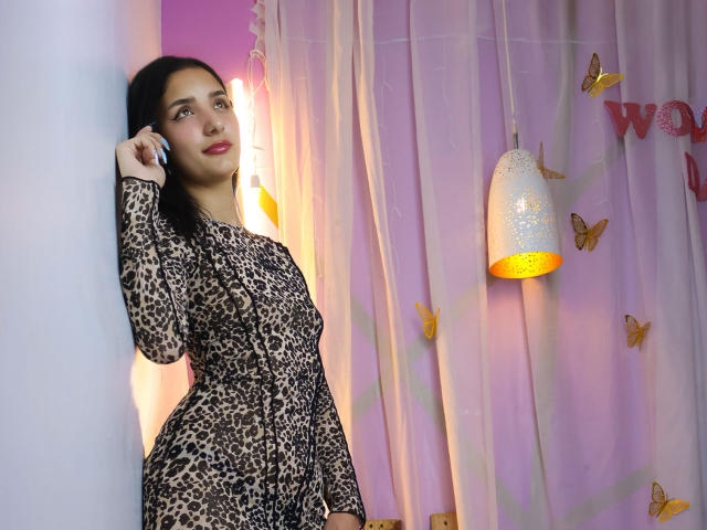 ArisSuarez staat leunend tegen een muur, gekleed in een strakke jurk met luipaardprint, en houdt een telefoon op haar oor. Een helder roze en oranje achtergrond, een hanglamp, gordijnen met een vlindermotief. Heel aantrekkelijk..