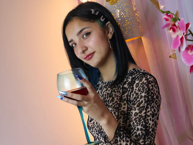 ArisSuarez, con cabello negro y piel de color marrón claro, sostiene un vaso con un líquido rojo, vistiendo una blusa con estampado de leopardo, y está mirando la cámara, con una ligera sonrisa. Flores rosadas en el fondo.