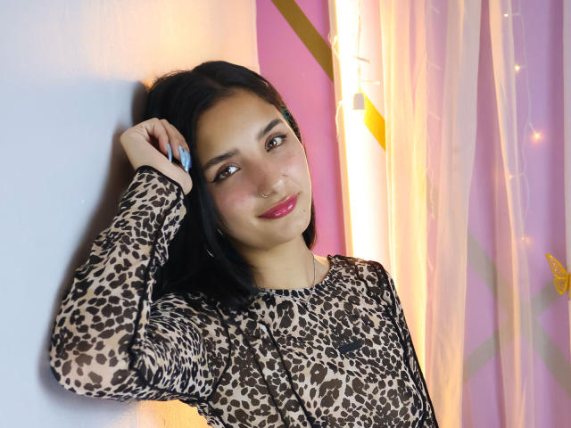 ArisSuarez, con la pelle marrone chiaro e i capelli scuri, indossa una maglietta a stampa leopardo, appoggiata a una parete azzurra, con una mano capelli, sorriso con labbra rosa, sfondo rosa e giallo chiaro..