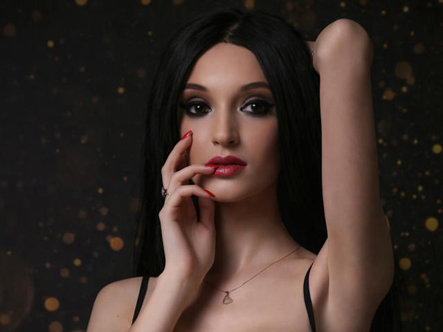 Fotografie von MaiaMoxie, einer dunkelhaarigen Frau mit rotem Lippenstift, schwarzem Eyeliner und rotem Nagellack, die ihren linken Arm hebt. Ihre Gesicht berührend, trägt sie ein schwarzes Top mit Trägern, dunkler Hintergrund mit Bokeh-Lichtern..