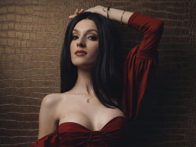 MaiaMoxie, uma mulher de pele clara com longos cabelos pretos, veste um vestido vermelho com um ombro descoberto, revelando sua região do colo. Ela está levantando o braço direito, com a mão no cabelo, olhando sedutivamente para a câmera..