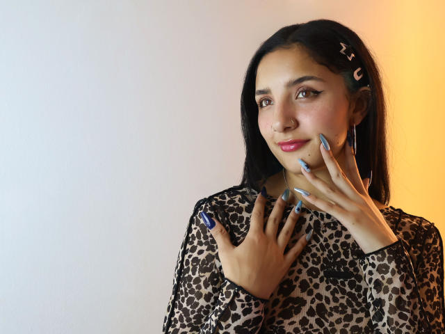 ArisSuarez, with dark hair and blue nails, touches her cheek with a hand over a leopard-print top, against a gradient white to yellow background. Fucking sexy, confident expression.