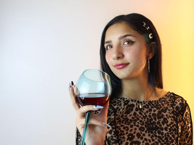 ArisSuarez houdt een glas rode wijn vast, en draagt een top met luipaardprint. Ze heeft lang zwart haar, roze lippenstift en een lichte glimlach. De achtergrond is een overgang van wit naar geel. De uitdrukking van ArisSuarez is aantrekkelijk..