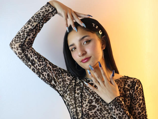 ArisSuarez, a sexy woman with black hair, wears a leopard-print top, poses seductively with one hand on her head and the other on her neck, blue nails, yellow-orange background.