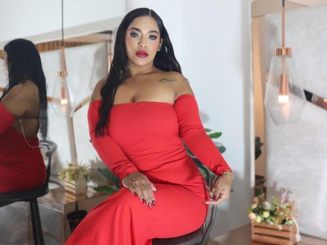 Lana Rossel, con cabello negro largo, vestido rojo con un hombro descubierto, sentada en una silla, con un tatuaje en el hombro izquierdo, sexy, mirada intensa. labios rojos, piel clara, ambiente moderno, plantas en el fondo, lámparas colgantes de cobre..