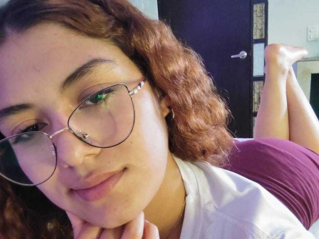 Close-up de TaniaLux, uma mulher curvilínea com cabelos castanhos cacheados, usando óculos e uma camiseta branca, com o queixo apoiado em... Mãos e pernas levantadas ao fundo. Uma vibe super relaxante..