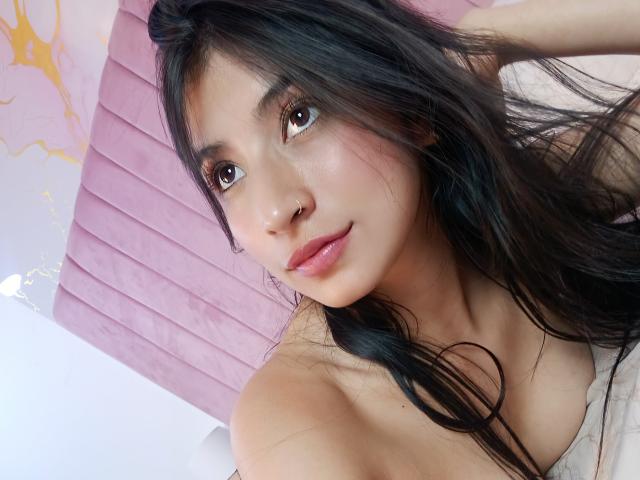 Primer plano de Naia Sanders, una joven con cabello largo y negro, piel clara y ojos marrones, mirando directamente a la cámara. con una sonrisa sutil. Tiene una figura delgada, labios rosados y una camiseta blanca. Su mano está en su cabello, creando una pose ligeramente provocativa..