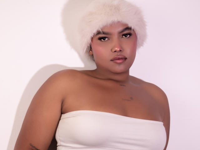 ChrystalFiore, con la pelle abbronzata, indossa un cappello di pelliccia bianca e un top bianco senza spalline, guardando direttamente l'obiettivo. Espressione neutra.
piccoli tatuaggi sul suo petto. Sfondo bianco. Incredibilmente sexy..