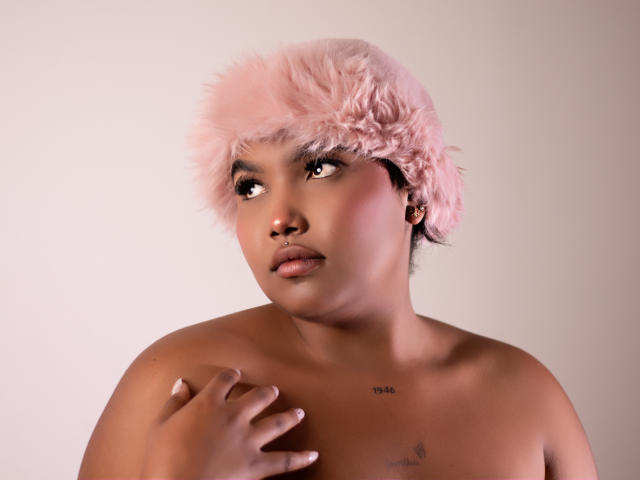 Photograph of ChrystalFiore, topless with short, fluffy pink hair, light brown skin, and a small tattoo on the right side of the chest. Hand on chest, intense gaze, slight smile.