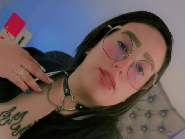 Close-up of NatalieHarris, wearing round glasses, black choker with heart ring, red lipstick, and black hair. Tattoos on neck, blue eye makeup, hand with ring, sexy, suggestive pose.